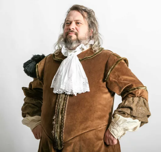 Man met een bruin historisch kostuum en een witte kraag, met lange haren en een zelfverzekerde houding.