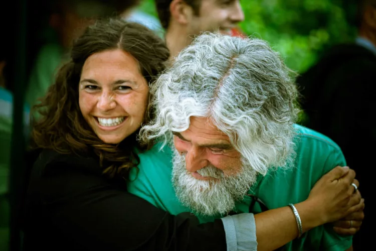 Vrouw met krullend haar omarmt een man met grijs haar en baard, beiden lachend.