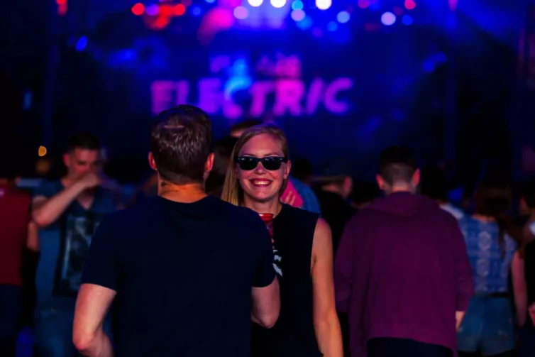 Vrouw met zonnebril en glimlach op het We Are Electric Festival 2015 met mensen op de achtergrond.
