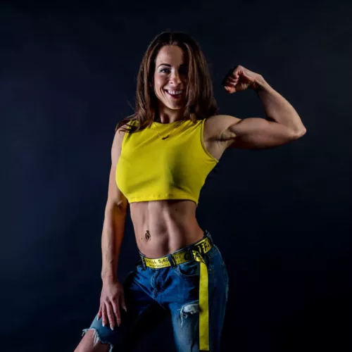 Vrouw in een gele crop top en blauwe jeans poseert met een gespierde arm.