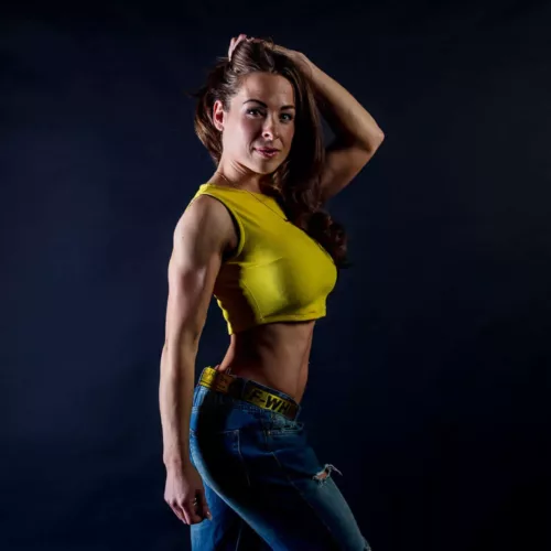 Vrouw in een gele crop top en spijkerbroek poseert met een hand in haar haar.