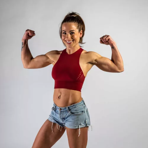 Vrouw met een rode sporttop en denim shorts poseert met gebalde vuisten en een glimlach.