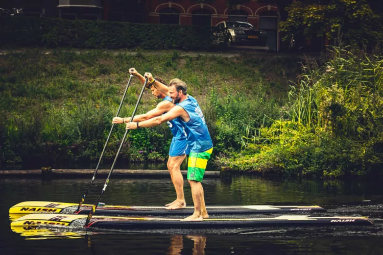 Twee mannen paddleboarden samen op een waterweg met een groene oever.