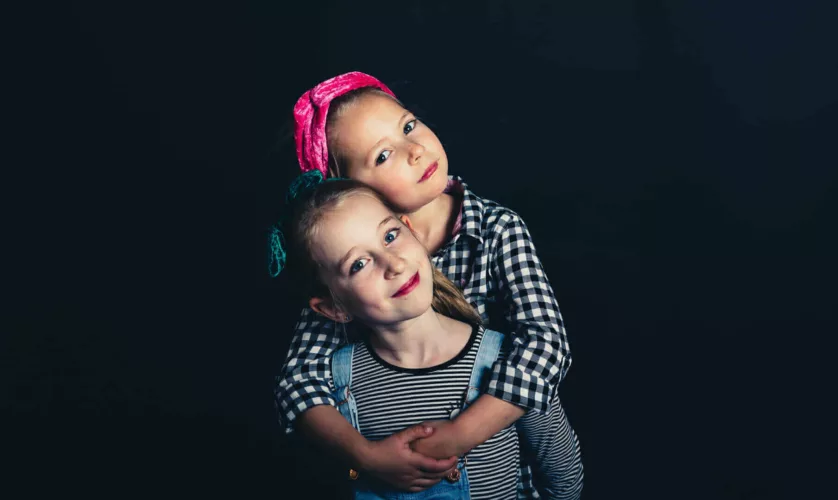 Twee meisjes poseren samen met een zwarte achtergrond, een met een haarband en de ander met een schort.