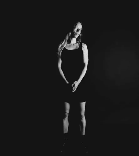 Zwart-wit portret van een vrouw met lange haren, gekleed in een zwarte jurk, staand met handen voor zich.