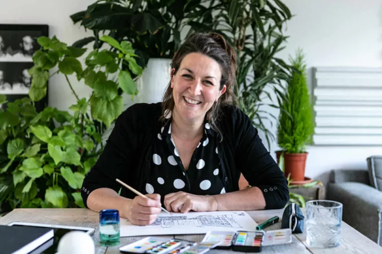 Vrouw met een pen en tekenmaterialen, zittend aan een tafel met planten op de achtergrond.