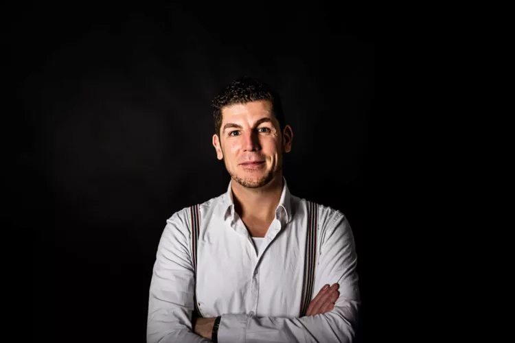 Portret van een man met een wit overhemd en bretels, staande met armen over elkaar.