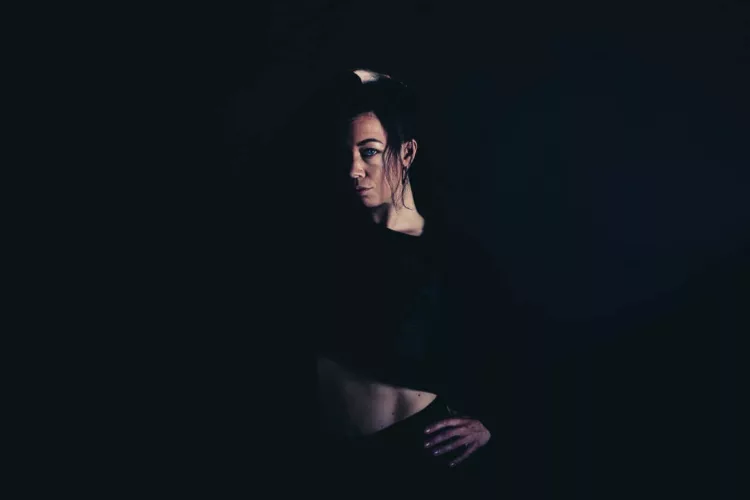 Portret van een vrouw met een hand op haar hoofd, gekleed in een zwarte outfit, in een donkere setting.