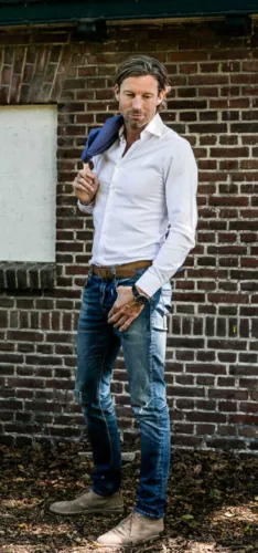 Man met een witte blouse, spijkerbroek en schoenen, poseert met een jas over zijn schouder.