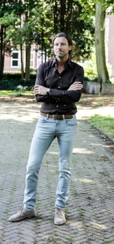 Man met lang haar in een donker shirt en lichte jeans, staand met zijn armen gekruist.
