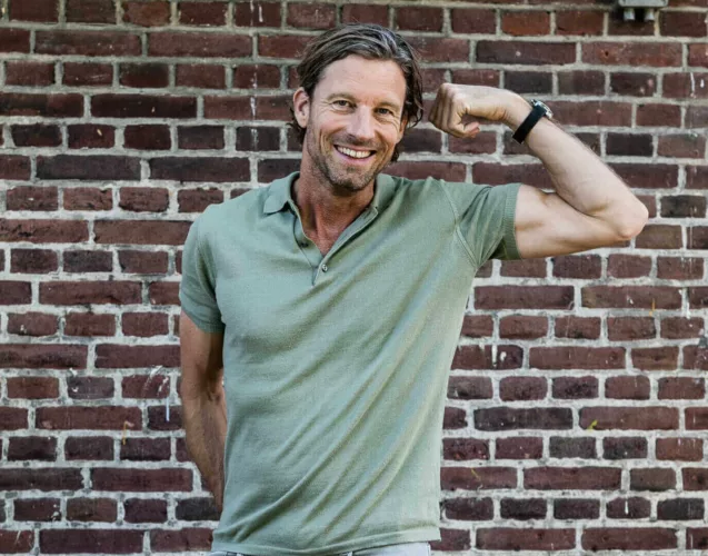 Man met een groene polo die zijn arm buigt en glimlacht voor een bakstenen muur.