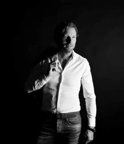 Man met een wit overhemd en een zwarte achtergrond, poseert met een hand op zijn schouder.