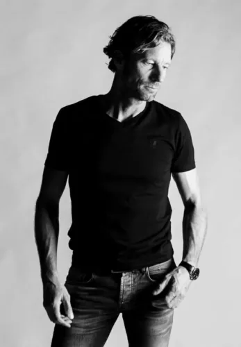 Man in zwart T-shirt en jeans poseert met een neutrale uitdrukking in zwart-wit fotografie.