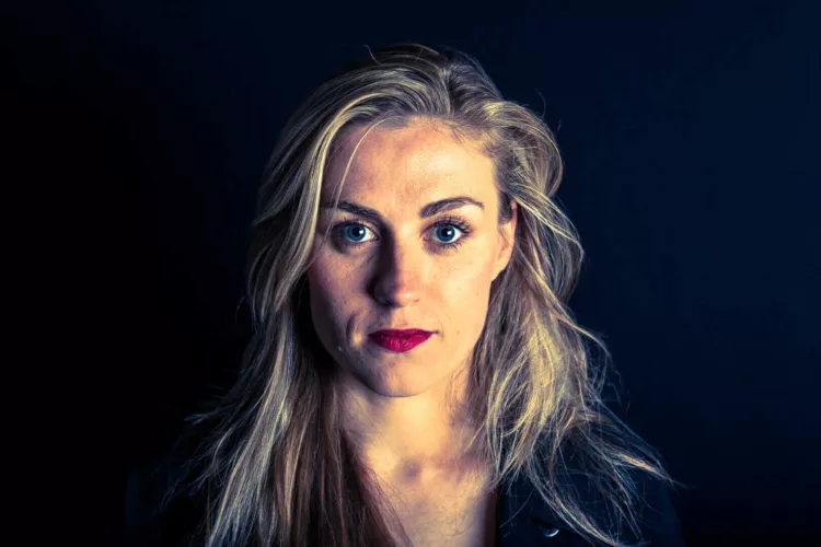 Portret van een vrouw met lange blonde haren en blauwe ogen, tegen een donkere achtergrond.