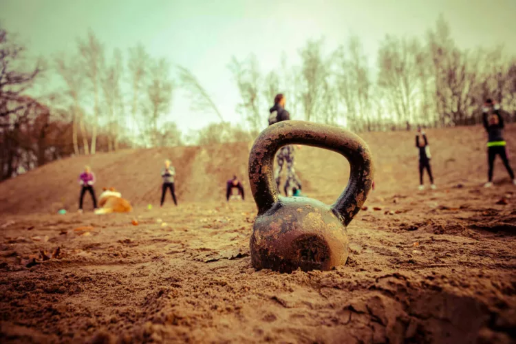 Een kettlebell ligt op de zandgrond terwijl mensen in de achtergrond aan een training deelnemen.