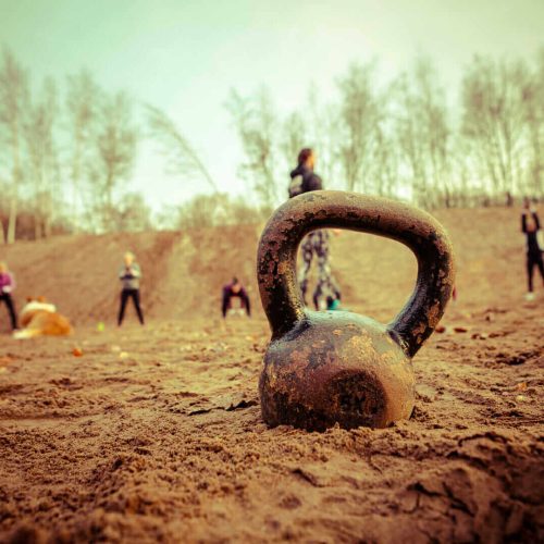 Een kettlebell ligt op de zandgrond terwijl mensen in de achtergrond aan een training deelnemen.