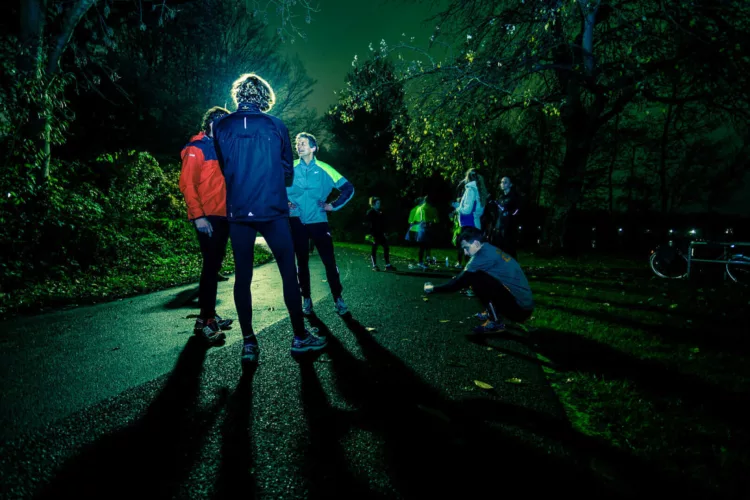 Een groep hardlopers in sportkleding staat op een pad tijdens de nacht met schaduwen op de grond.