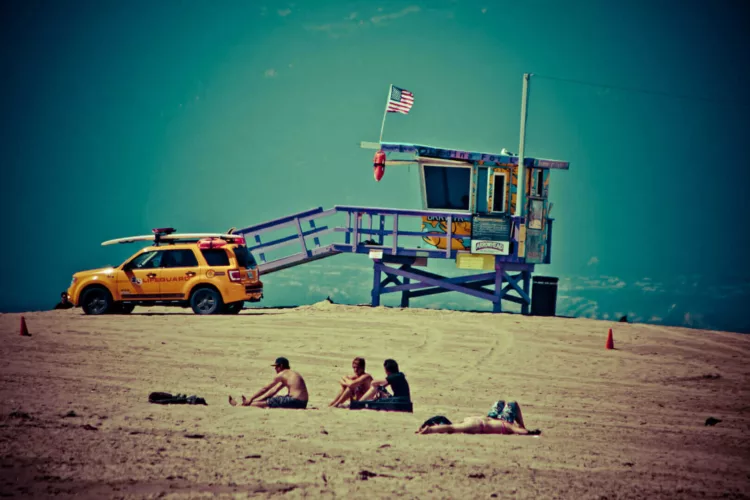 Een strandwachtpost met een gele auto en mensen die op het zand zitten.