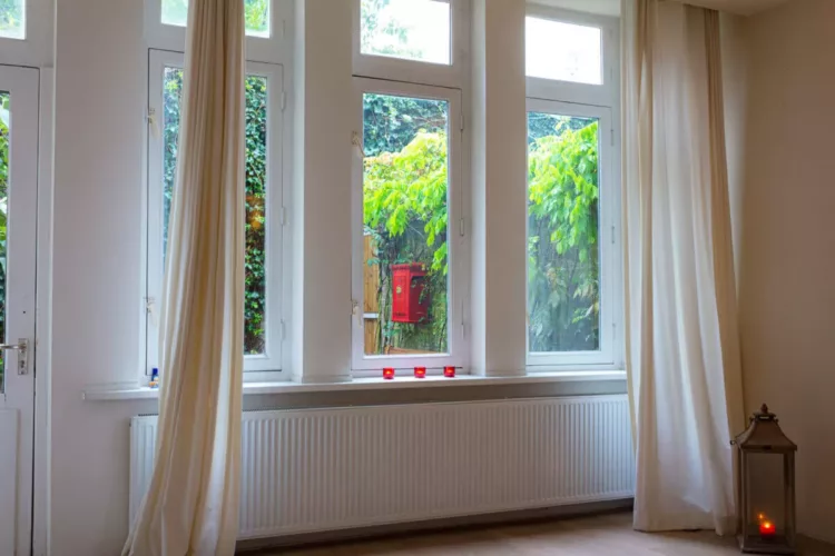 Raam met witte gordijnen en een verwarmingselement onder het raam in een lichte ruimte.