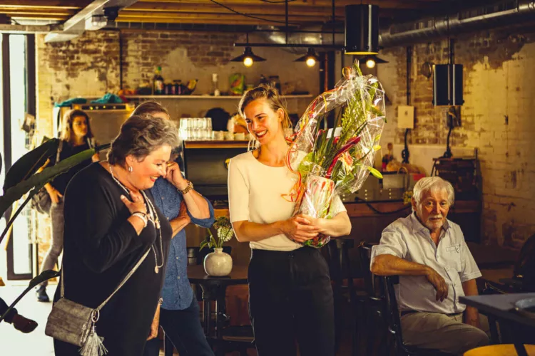 Vrouw met een bloemenboeket in een café, omringd door andere mensen.