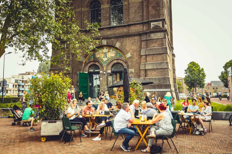 Groep mensen die aan tafels buiten zitten bij een eetgelegenheid met een gebouw op de achtergrond.