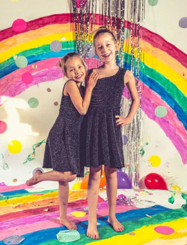Twee meisjes in zwarte jurken staan voor een kleurrijke regenboogachtergrond met ballonnen en confetti.