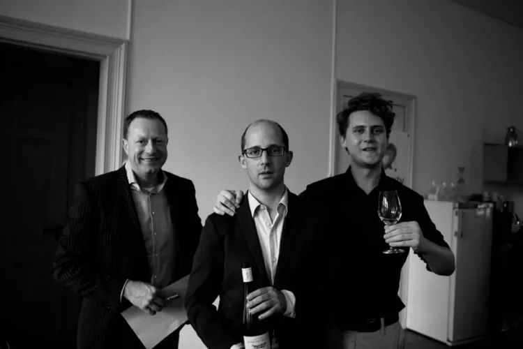 Drie mannen poseren samen met een wijnfles en een glas in zwart-wit foto.
