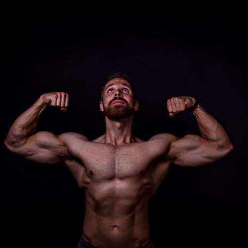 Man met een baard toont zijn spieren met gebogen armen tegen een zwarte achtergrond.
