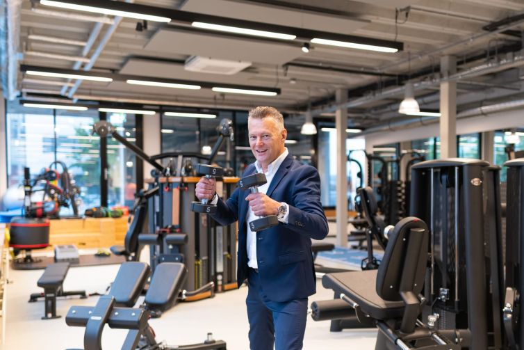 Man in een pak die twee dumbbells vasthoudt in een moderne fitnessruimte met apparatuur op de achtergrond.