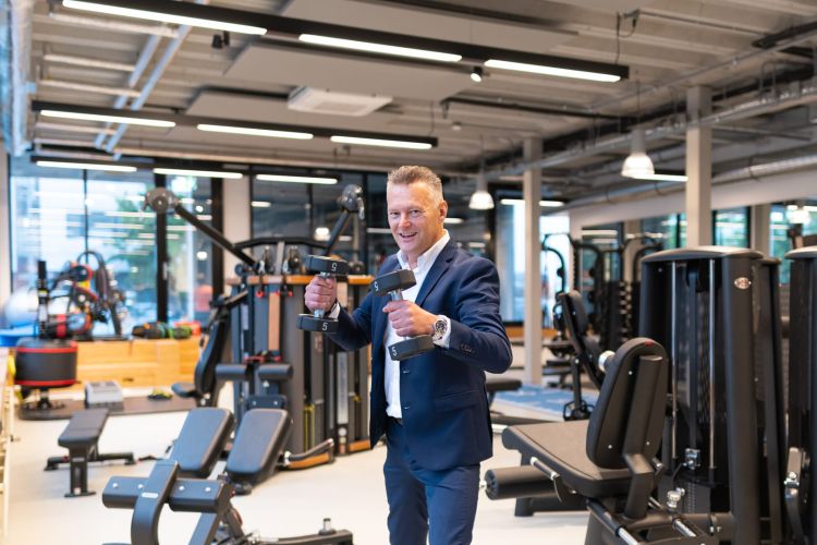 Man in een pak die twee dumbbells vasthoudt in een moderne fitnessruimte met apparatuur op de achtergrond.