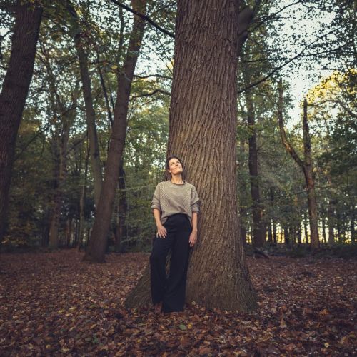 Vrouw in een grijze trui leunt tegen een grote boom in een bos met bladeren op de grond.