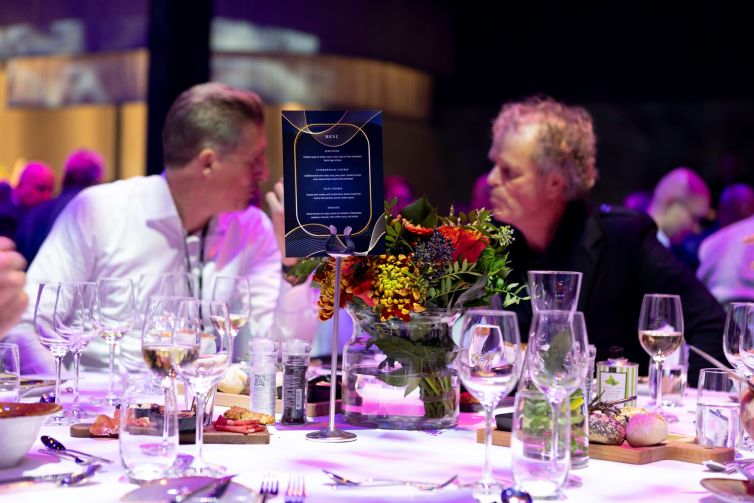 Twee mannen in gesprek aan een gedekte tafel met bloemen en glazen.