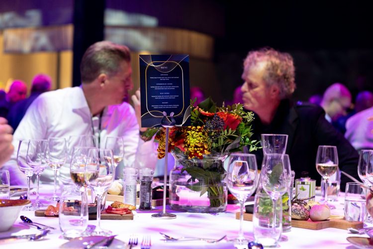 Twee mannen in gesprek aan een gedekte tafel met bloemen en glazen.