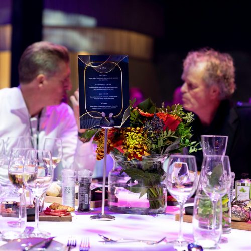 Twee mannen in gesprek aan een gedekte tafel met bloemen en glazen.