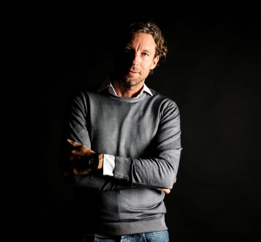 Man met een grijze trui en een witte blouse zichtbaar onder de trui, met gekruiste armen.